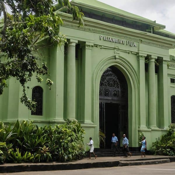 Green Fiji Parliament building with people walking outside, lush greenery around.
